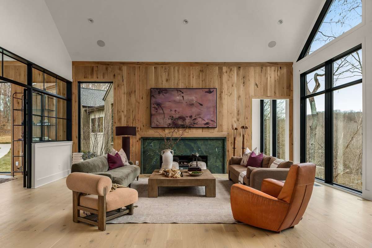 Living room featuring soaring vaulted ceilings, a statement fireplace, and expansive windows that fill the space with natural light while framing peaceful wooded views.