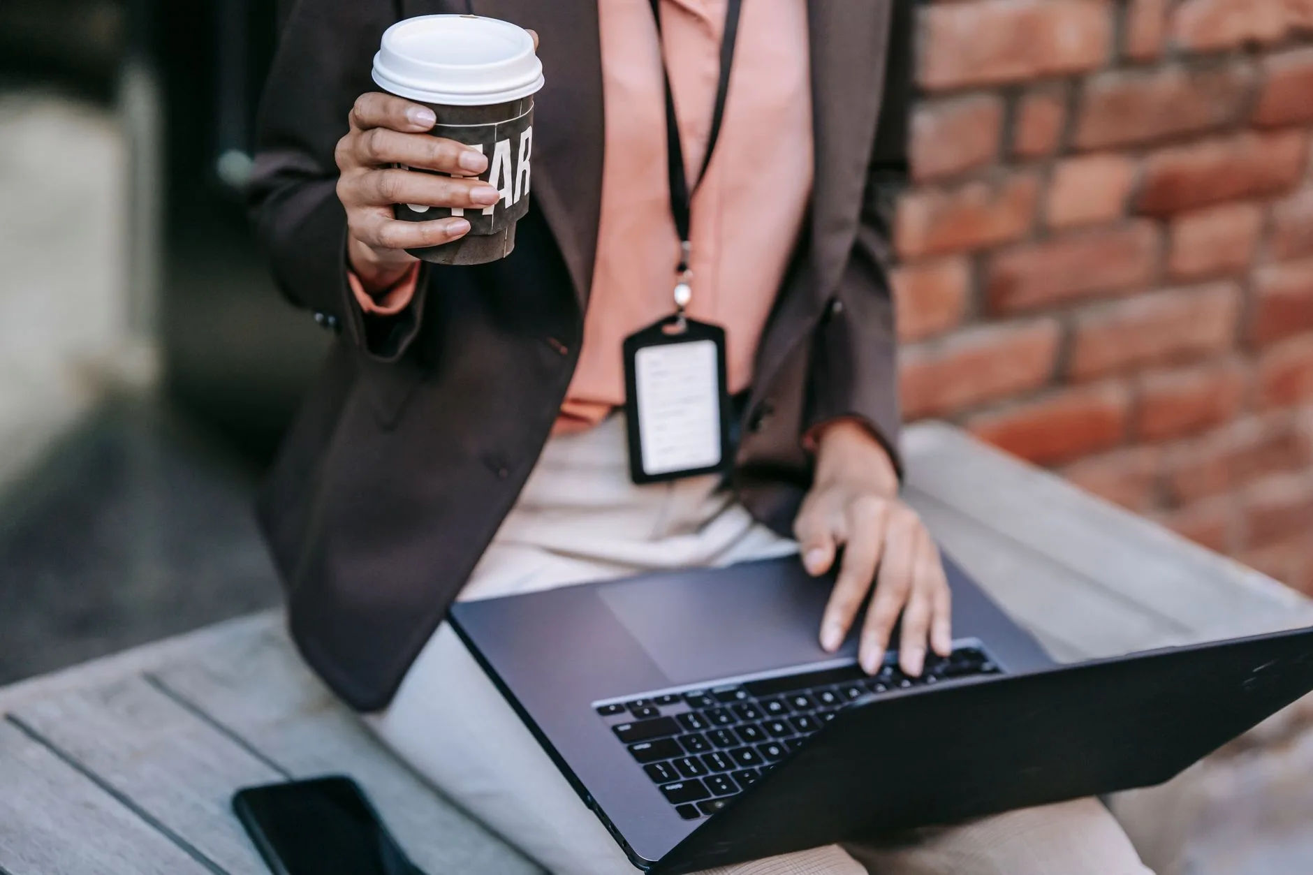 Crop anonymous female entrepreneur wearing elegant black jacket with badge drinking coffee while using laptop sitting on bench