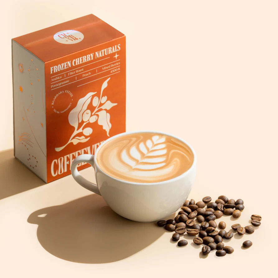 Cup of coffee with latte art, coffee beans, and a box labeled 'Frozen Cherry Naturals Coffee' on a beige background.