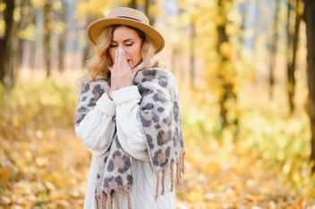 A blonde woman wearing a hat and blowing her nose in an autumn background