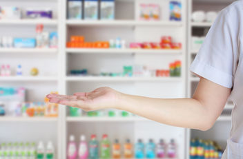 Imagen de la mano de un farmacéutico frente a los estantes de la farmacia.