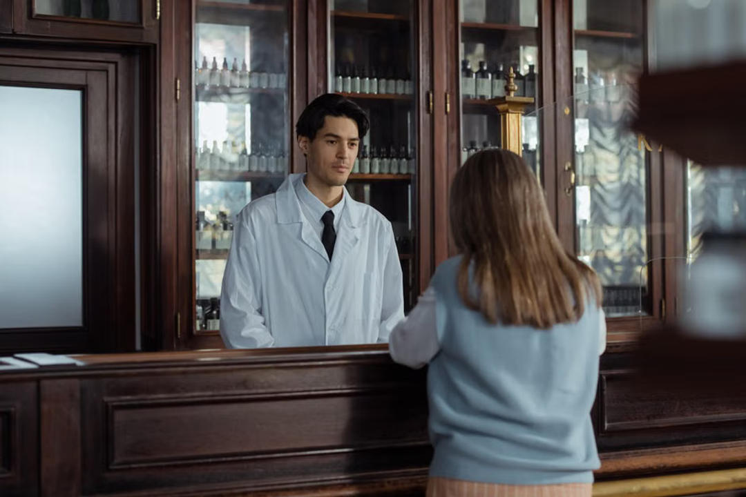 File image: Man in white coat (pharmacist) consulting with woman at pharmacy counter with product cabinets behind.