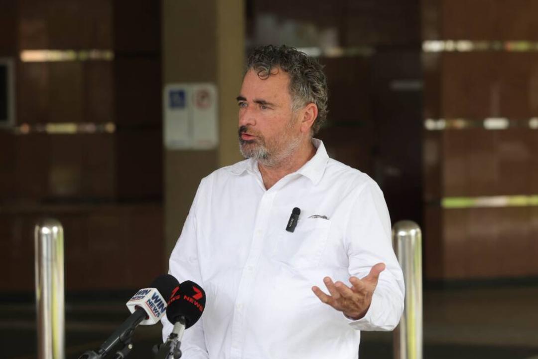 Jason Gordon. Man in white shirt speaking at microphones at formal government building setting