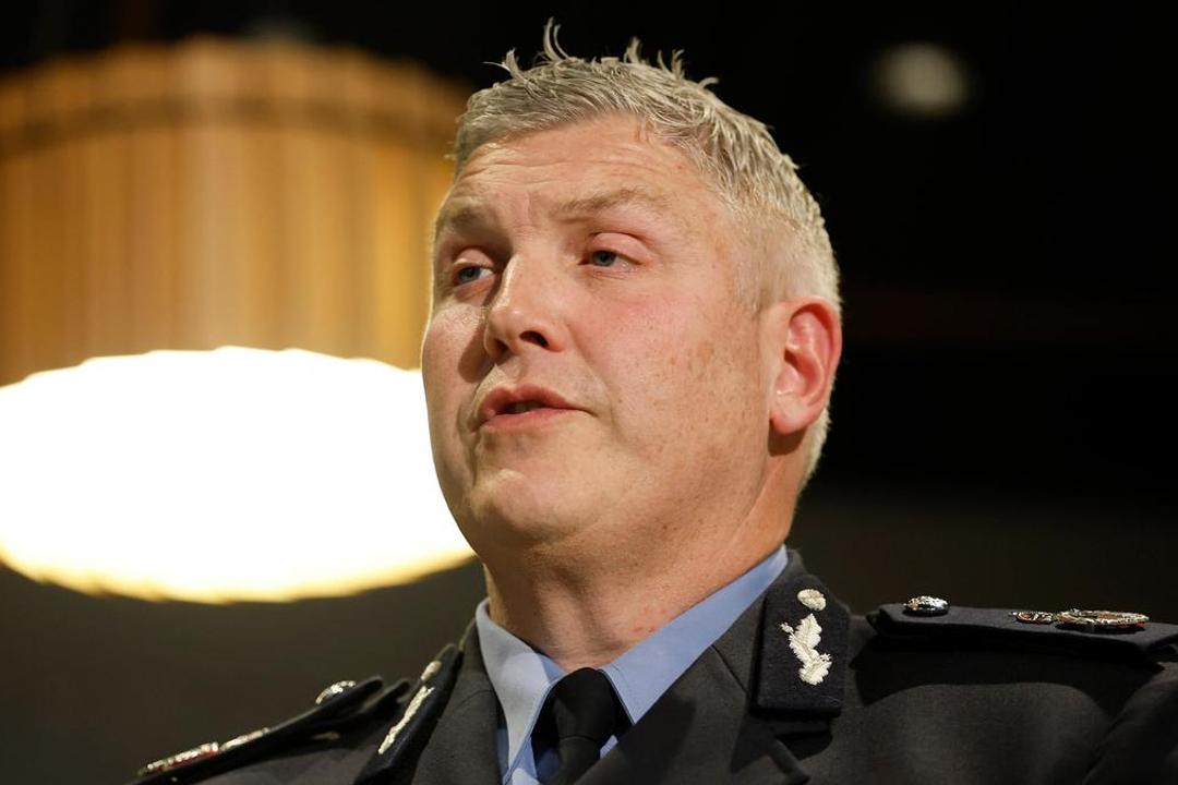 Man in dark police uniform with light blue shirt speaking indoors under lamp
