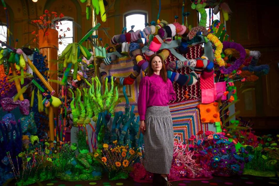 Woman in magenta sweater stands amid colorful hanging sculptural art installation in indoor gallery space.
