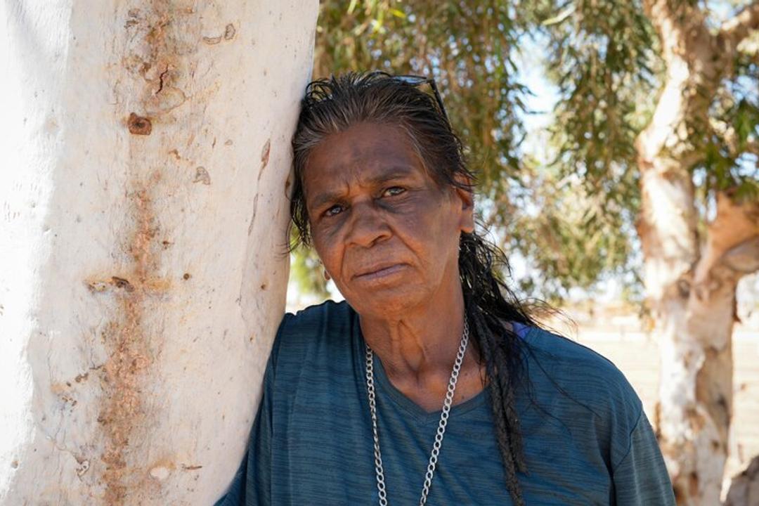 Middle-aged woman in dark shirt leaning against weathered wall with eucalyptus trees behind
