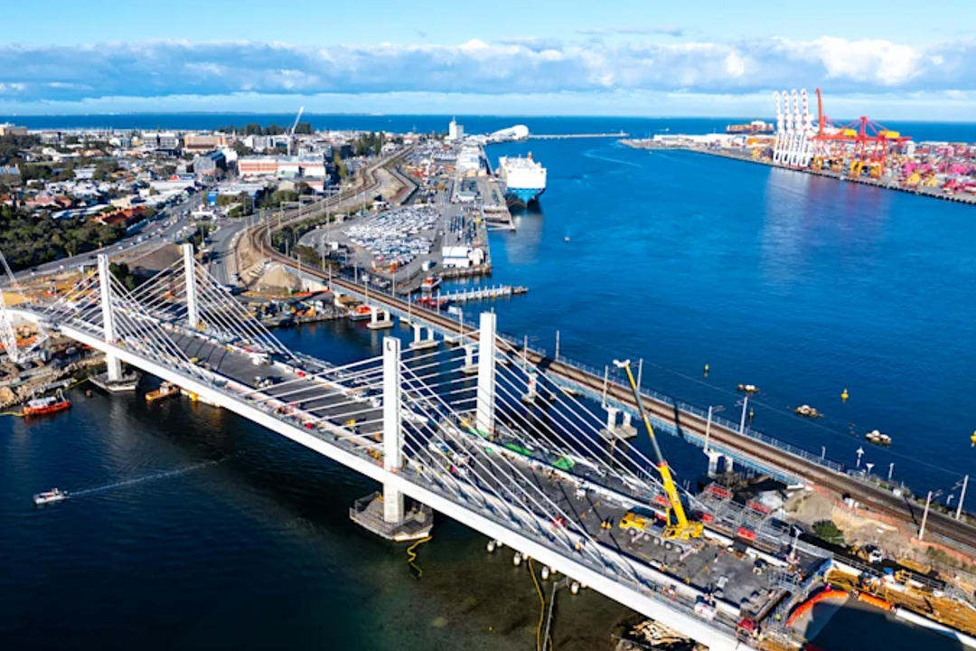 Aerial view of cable-stayed bridge with white pylons spanning blue water, harbor with ships and urban development visible