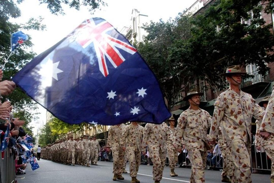 Australian military personnel in desert camouflage uniforms holding large Australian flag at urban public event