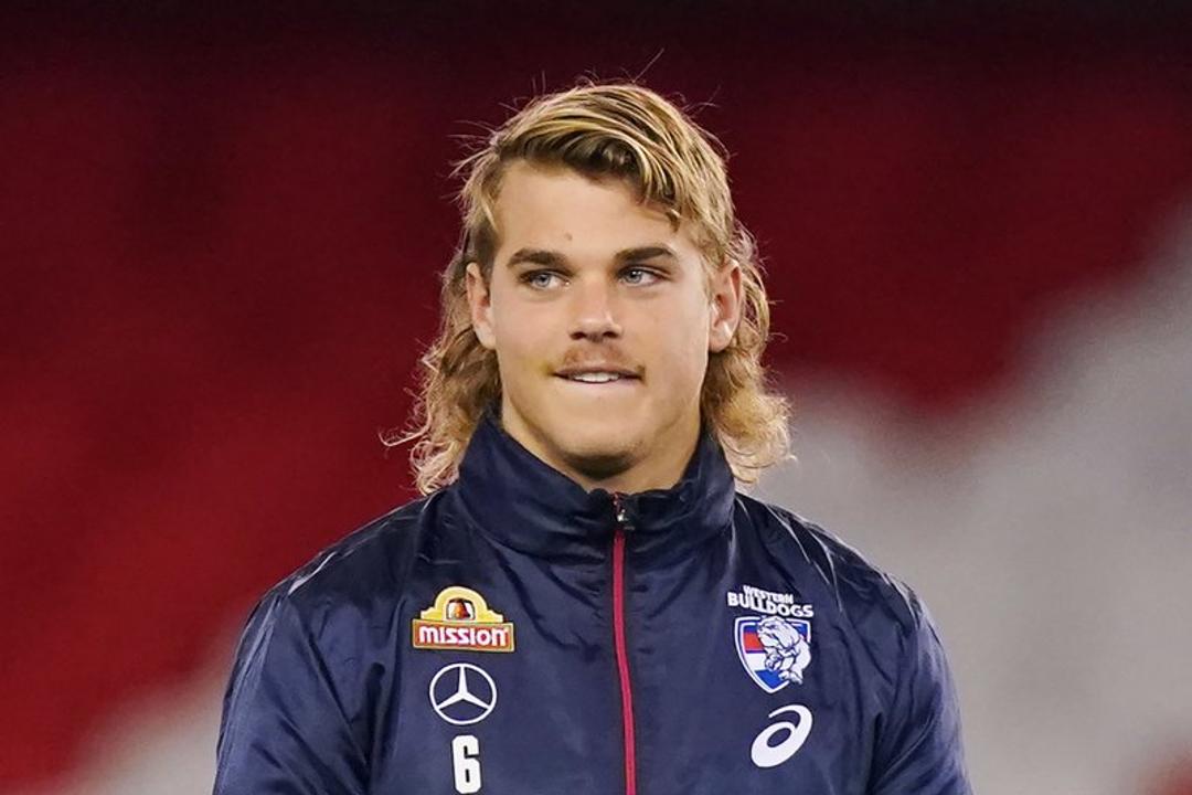 Bailey Smith. A young man with shoulder-length blonde hair wearing a navy Western Bulldogs AFL training jacket bearing the number 6, holding a yellow Sherrin football.
