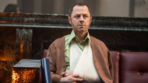 Man seated indoors wearing brown cardigan over green shirt, serious expression.