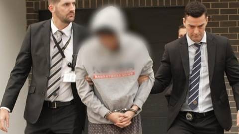 Man in hoodie escorted by two officials in business suits outside brick building