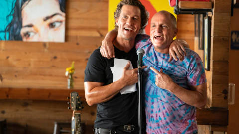 Two men smiling at a microphone in a wooden venue with a guitar visible, apparently at a promotional event