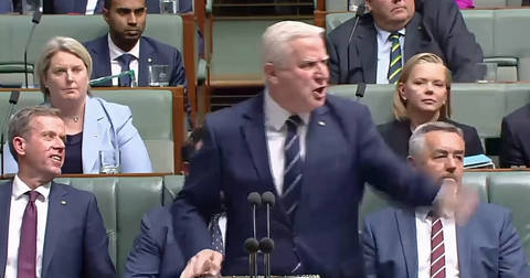 A man in dark suit speaking at parliamentary dispatch box with multiple senators seated behind him