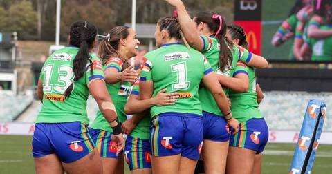 Female rugby league players in green and blue Raiders uniforms with rainbow armbands, celebrating together on field