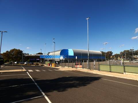 Contemporary curved-roof sports arena building with outdoor courts and lighting infrastructure.