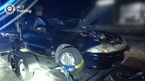 Towed dark vehicle with open door on flatbed at night, second vehicle visible, ACT Police branding
