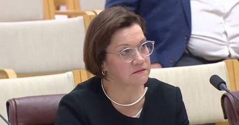 Woman wearing glasses and black blazer at formal government committee meeting with microphone