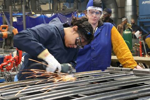 Apprentice welding metal under supervision, sparks flying, industrial workshop setting.