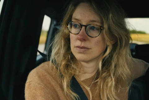 Nathan Fielder. Woman wearing glasses and tan cardigan sitting in car, looking to side with serious expression