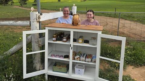 Two people at white cabinet roadside pantry stand with jars, goods and baked bread displayed