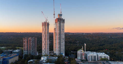 Two residential apartment towers under construction at dusk with construction cranes visible against sunset sky