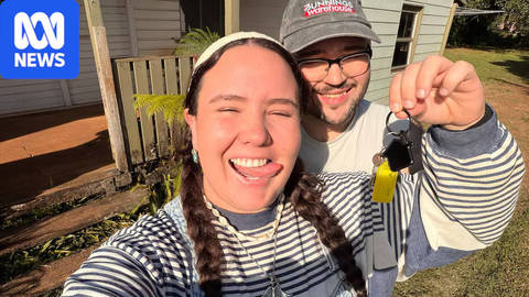 Two people holding house key outside suburban home, smiling at camera