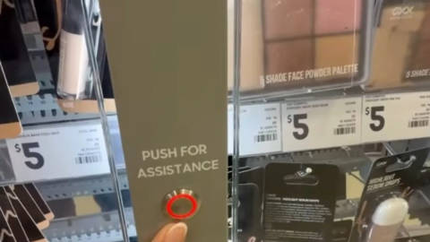Hand pointing at red assistance button on locked glass cabinet containing $5 makeup palette in retail display
