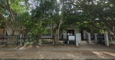 Heritage stone building with mature trees, metal fence, institutional setting
