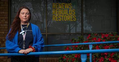 Woman in blue cardigan wearing staff lanyard standing at railing with 'Renews Rebuilds Restores' signage behind