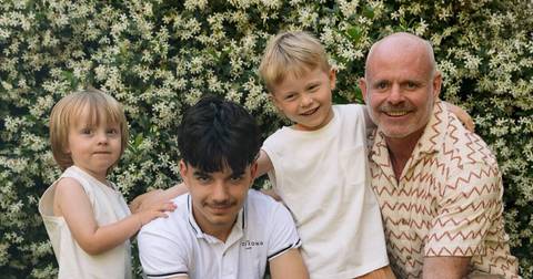 Four children and an adult man smiling outdoors against flowering shrubs
