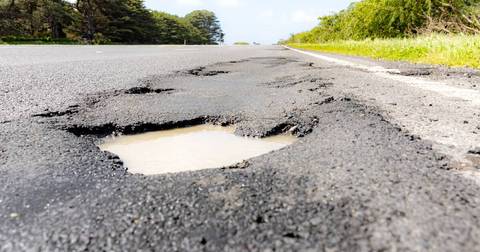Pothole filled with water in damaged asphalt road, grass verges visible beyond