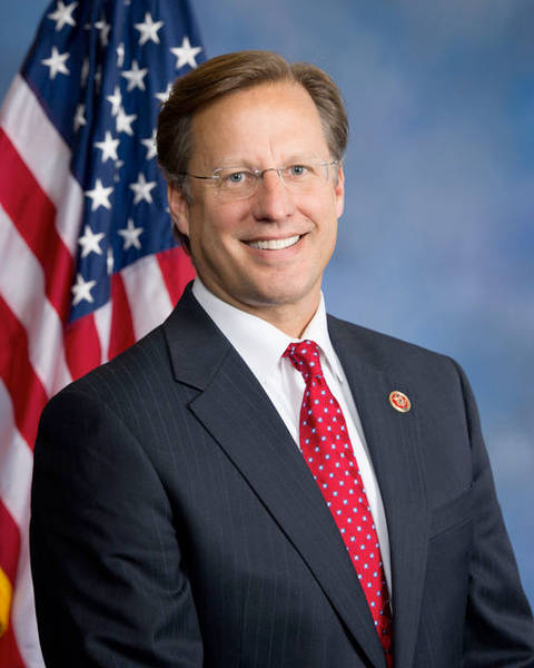 David Brat. Man in dark suit and red tie smiling before American flag, formal official portrait.