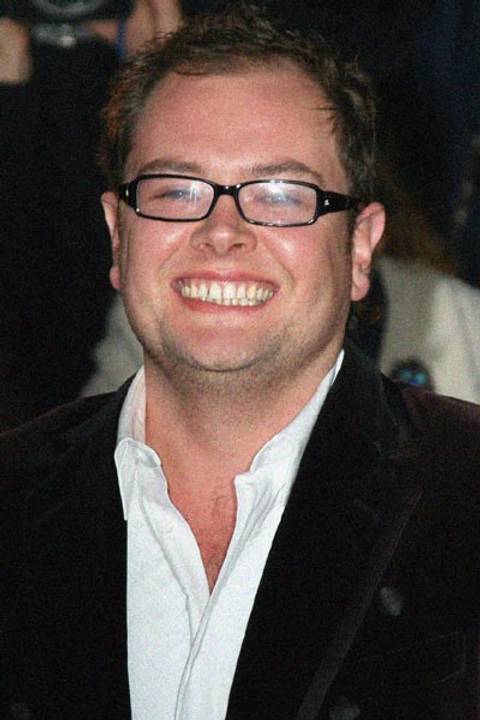 Alan Carr. A man wearing black-framed glasses and a dark velvet blazer over an open-collar white shirt, grinning broadly at the camera against a dark background at what appears to be a public