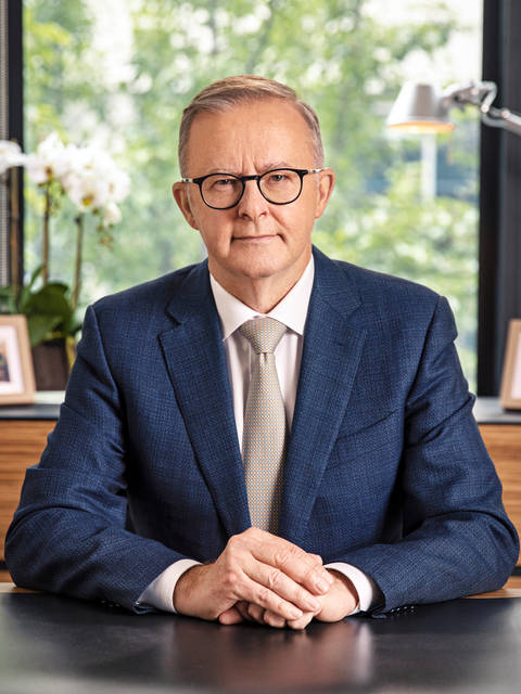 Anthony Albanese. A man wearing black-rimmed glasses and a navy blue suit with a light patterned tie, seated in front of a large window with green foliage and a desk lamp visible in the background.