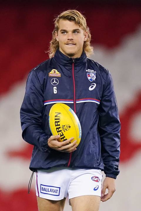 Bailey Smith. A young man with shoulder-length blonde hair wearing a navy Western Bulldogs AFL training jacket bearing the number 6, holding a yellow Sherrin football.
