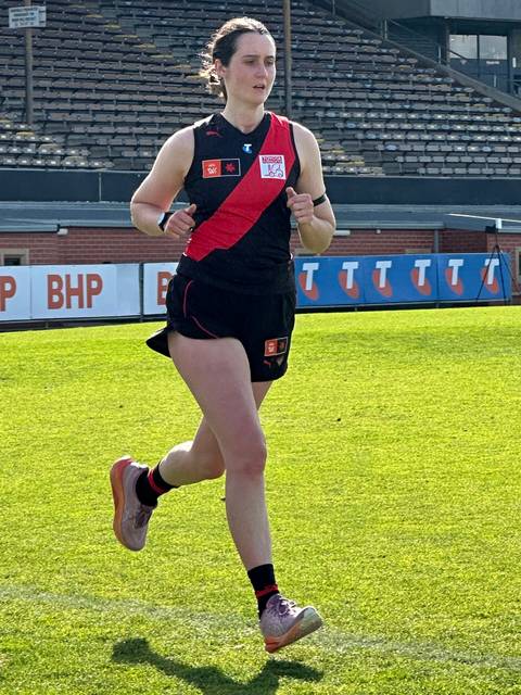 Bess Keaney. A woman wearing a black and red Australian rules football uniform runs across a grass oval with stadium seating and sponsor hoardings visible in the background.