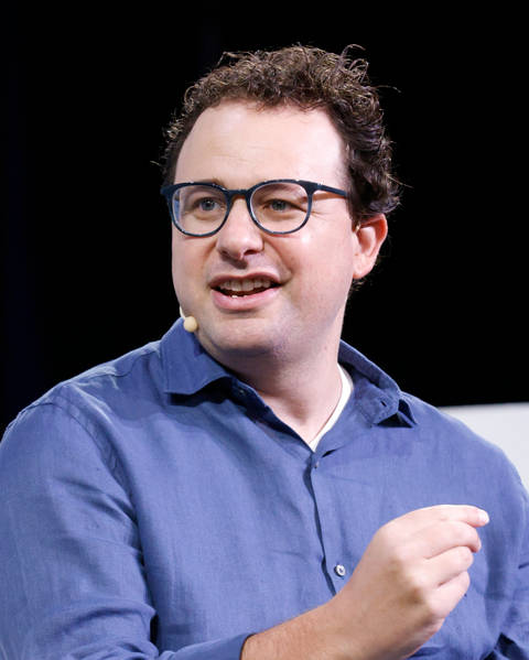 Dario Amodei. A man with curly dark hair and dark-framed glasses, wearing a blue linen shirt and a small earpiece microphone, speaks at what appears to be a public event against a dark backgroun