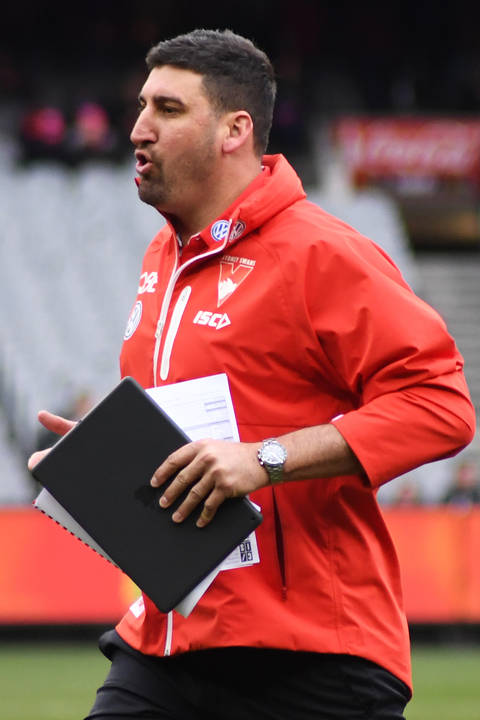 A man wearing a red Sydney Swans staff jacket holds a tablet and papers while walking purposefully on an AFL ground.