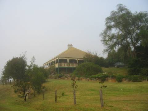 A colonial-style homestead with a wraparound veranda and pyramidal roof sits on a terraced hilltop surrounded by trees in misty conditions in the Hunter Valley.