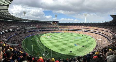 A panoramic view of the Melbourne Cricket Ground packed with tens of thousands of spectators during an AFL match, showing the oval green field, tiered grandstands wrapping the full