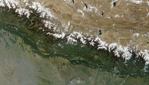 A satellite view of the Himalayan mountain range showing snow-capped peaks, green forested slopes, and the flat plains of the Indian subcontinent to the south with the Tibetan plat