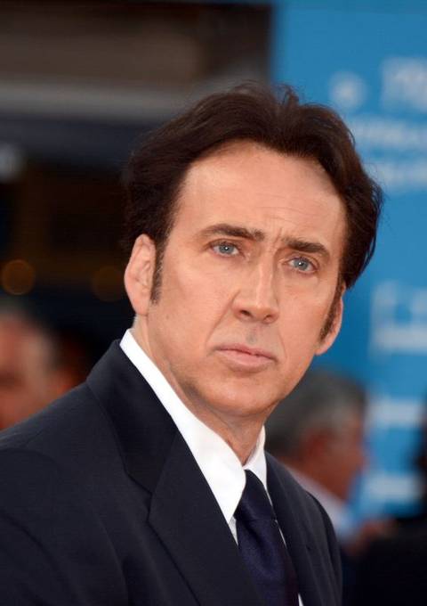 Nicolas Cage. A man with dark hair and blue eyes wearing a dark suit and tie, photographed outdoors at what appears to be a formal event with a blurred teal backdrop behind him.