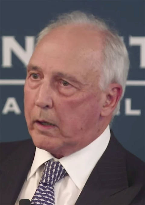 Close-up of an older man with grey hair wearing a dark suit and white shirt, speaking at what appears to be a press conference or official event.