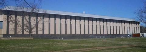 The exterior of the Royal Australian Mint, a long low-rise beige building with regularly spaced dark vertical pillar elements and a metal roof, photographed from across a grass law