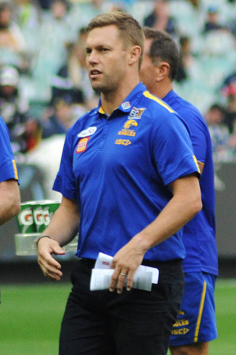 Sam Mitchell. A man wearing a blue and gold West Coast Eagles staff polo shirt walks across a football oval during a match, holding papers in one hand, with other team staff visible behind him.