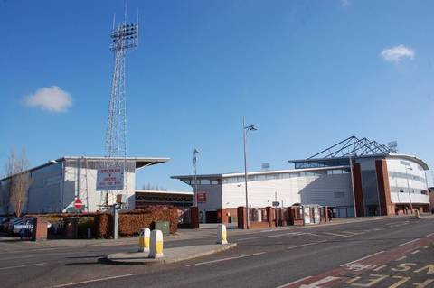 Exterior view of Wrexham AFC's Racecourse Ground on a clear day, with a floodlight pylon on the left and a matchday sign reading 'Wrexham v Chester' visible on the stand facade.