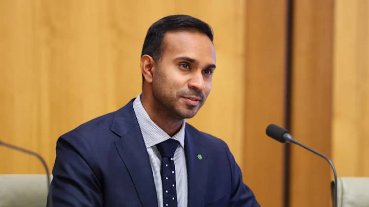 Man in navy suit and striped shirt speaking at microphone indoors against golden wall