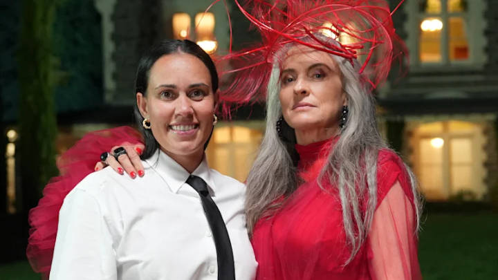 Two women at formal outdoor event, one in white shirt and tie, one in red outfit with red fascinator, building visible behind