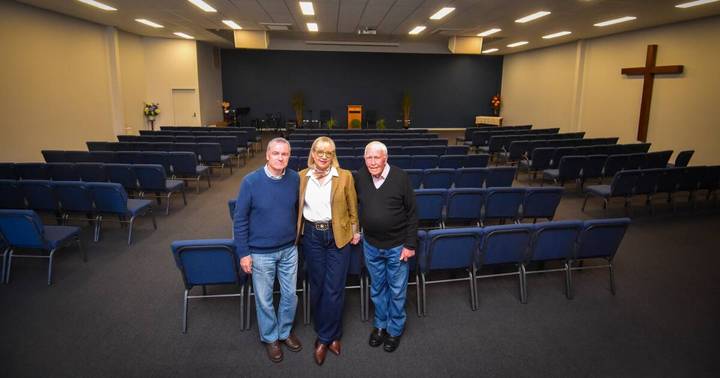 Three people in casual business wear pose in modern auditorium with blue seating
