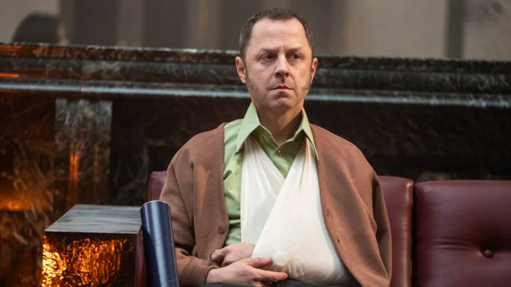Man seated indoors wearing brown cardigan over green shirt, serious expression.